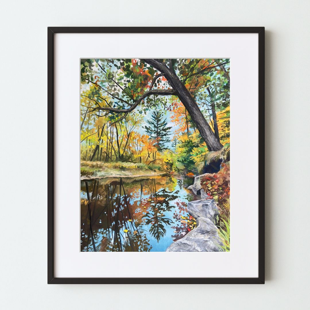 A mockup of a landscape art print in a black frame with a white mat. A still, reflective river runs through a forest during peak fall foliage. A tree near the viewer on the right leans out over the river, its branches overhead.