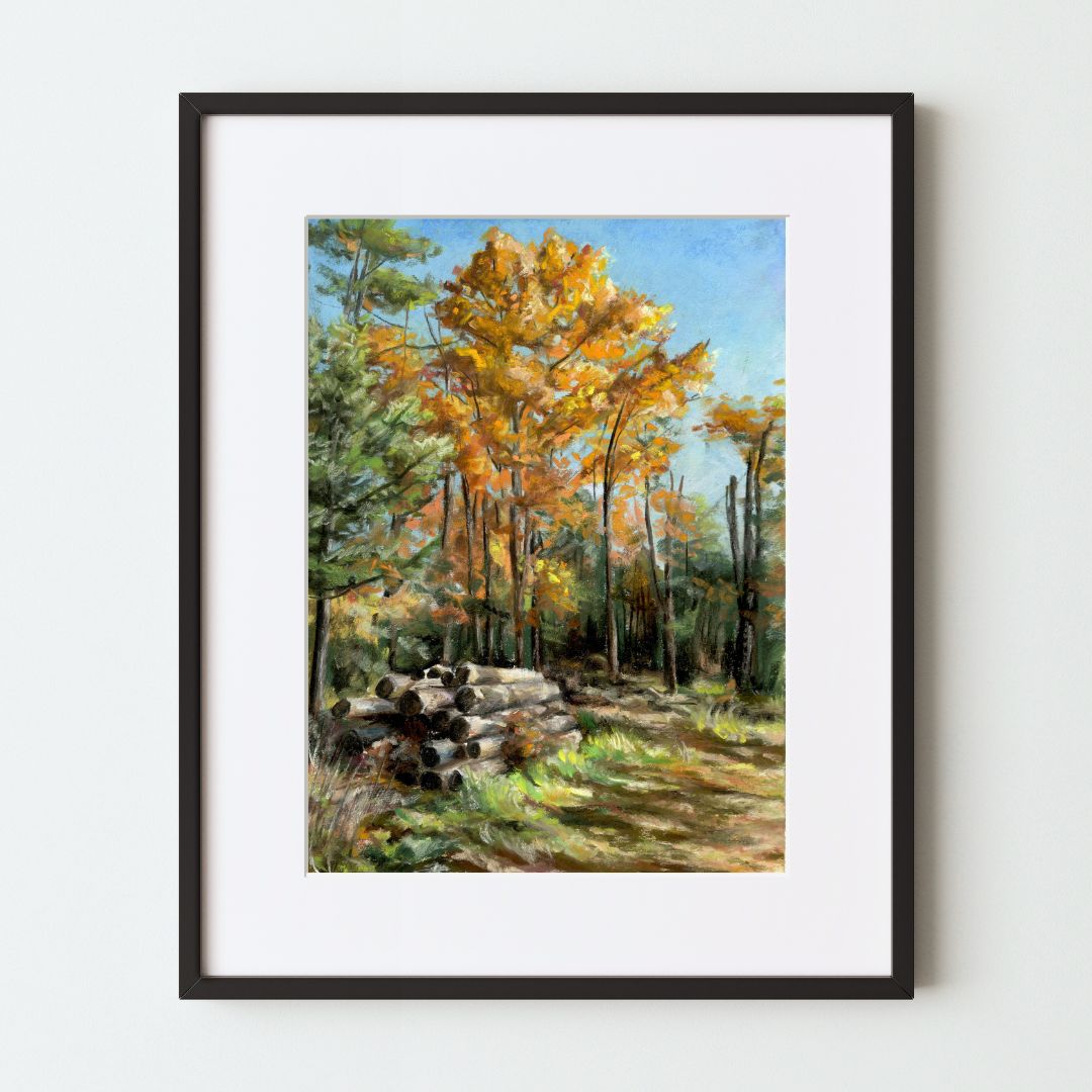 A mockup of a landscape art print in a black frame with a white mat. the painting shows several trees, some with fall colors, and a pile of logs next to a grassy path. The main colors in this nature scene are orange, blue and green.