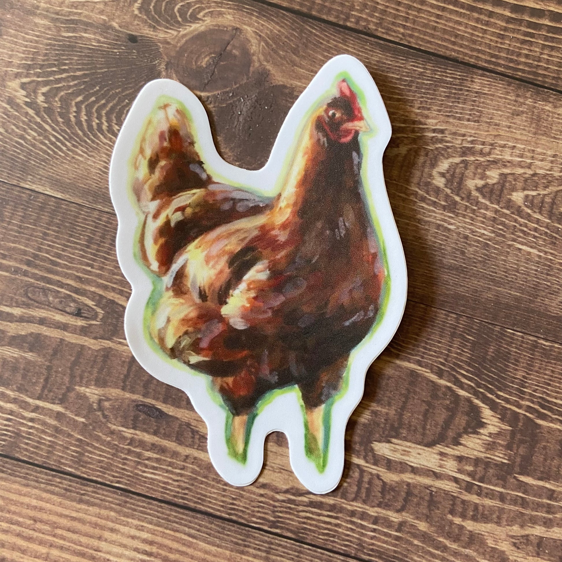 A chicken sticker sitting on a wooden surface. The decal shows a painting of an orange chicken in sunlight, and with both legs partially obscured by grass. The sticker has a thin white border and is die cut to shape.