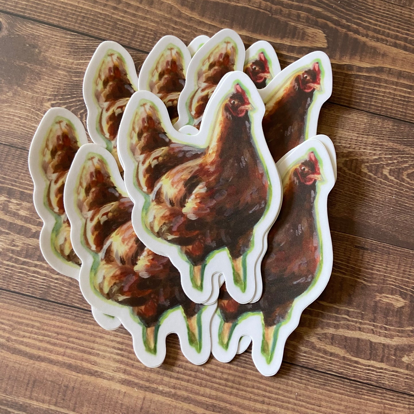 A small pile of vinyl stickers on a wood surface, showing a painted chicken standing in the sunlight. Each decal has a thin white border and is die-cut to shape.