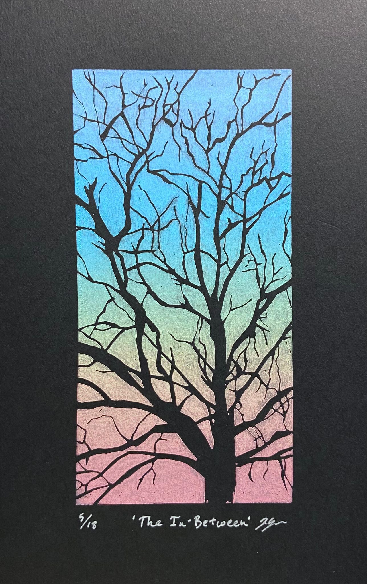 A 3x6 rainbow colored linocut relief print on black paper, showing a tree with bare branches. The tree is carved away to reveal the black underneath, and the printed area is the negative space between branches. Print is numbered, signed and titled.