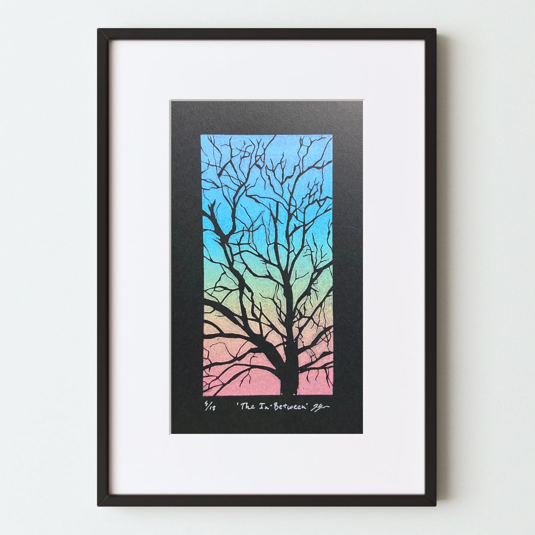 A mockup of a rainbow-colored tree branch art print in a black frame with a white mat. The linocut relief print on black paper shows a tree with bare branches carved away. The negative space between branches is printed with a rainbow roll technique, leaving the branches unprinted and black as the paper. 