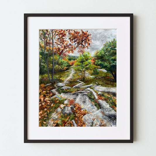 A mockup of a landscape art print in a black frame with a white mat. The print shows a gray rocky path through woods with green and brown foliage under an overcast sky.