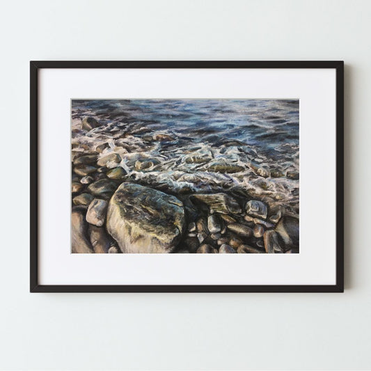 A mockup of an ocean-themed art print in a black frame with a white mat. The painting is a close look at some blue waves rushing over gray rocks on the beach. The focus is the largest rock, at the bottom and slightly left. 