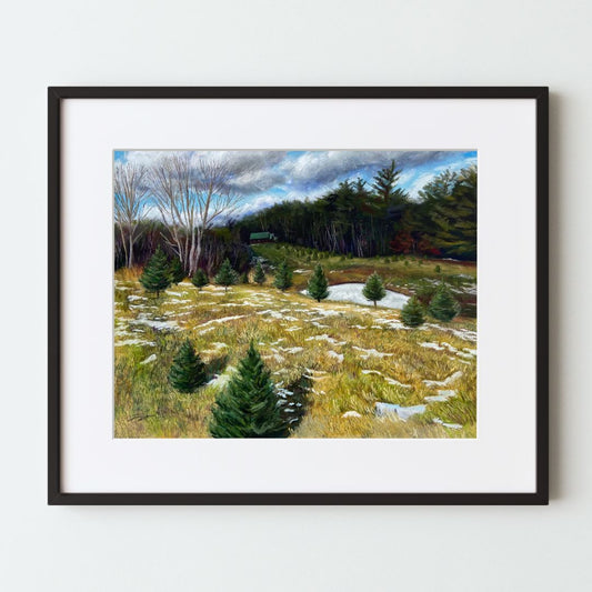 A mockup of a landscape art print in a black frame with a white mat. The painting shows a hill in the foreground with yellow grass, Christmas trees and patchy snow. Farther away is a snowy pond, more planted trees and the forest, and a far cabin.