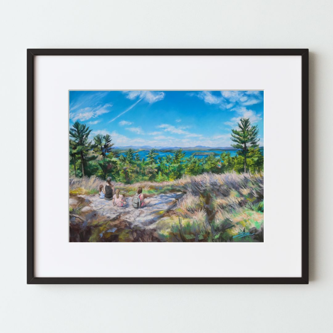 A mockup of a landscape art print in a black frame with a white mat. The painting shows a family of hikers on a rocky hill, looking out past a midground of green trees at Lake Winnipesaukee under a wide blue sky with sparse white clouds.